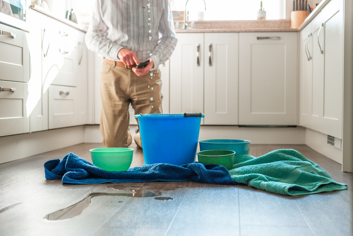 A senior man looks worried as he deals with a water leak in the kitchen ceiling. Buckets and pots are on the floor to catch the flowing water, and towels are soaking up the puddles. The man is defocused in the background, using his smart phone.